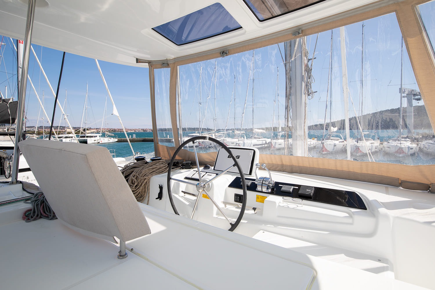Helm station of the Lagoon 46 with stainless steering wheel, B&G chartplotter, winches and clear cockpit enclosure looking out over the marina