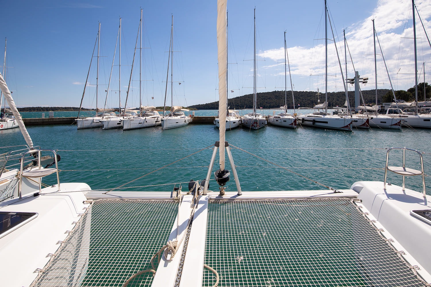 Trampoline netting between the two bows, looking out over moored yachts and the wooded shoreline of the marina.