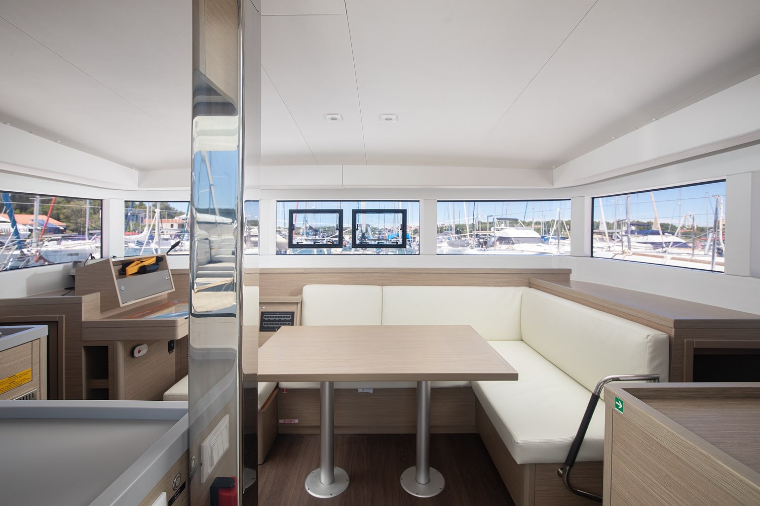 Saloon interior with a cream L-shaped settee, dining table and wraparound windows looking out over the marina