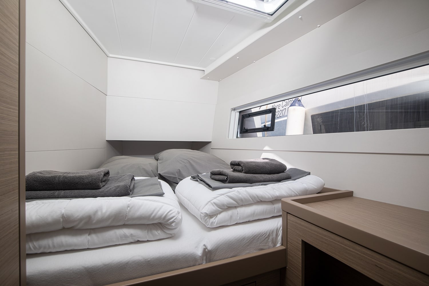 Double cabin with a made-up berth and towels, a long hull window and a shelf running along the bulkhead