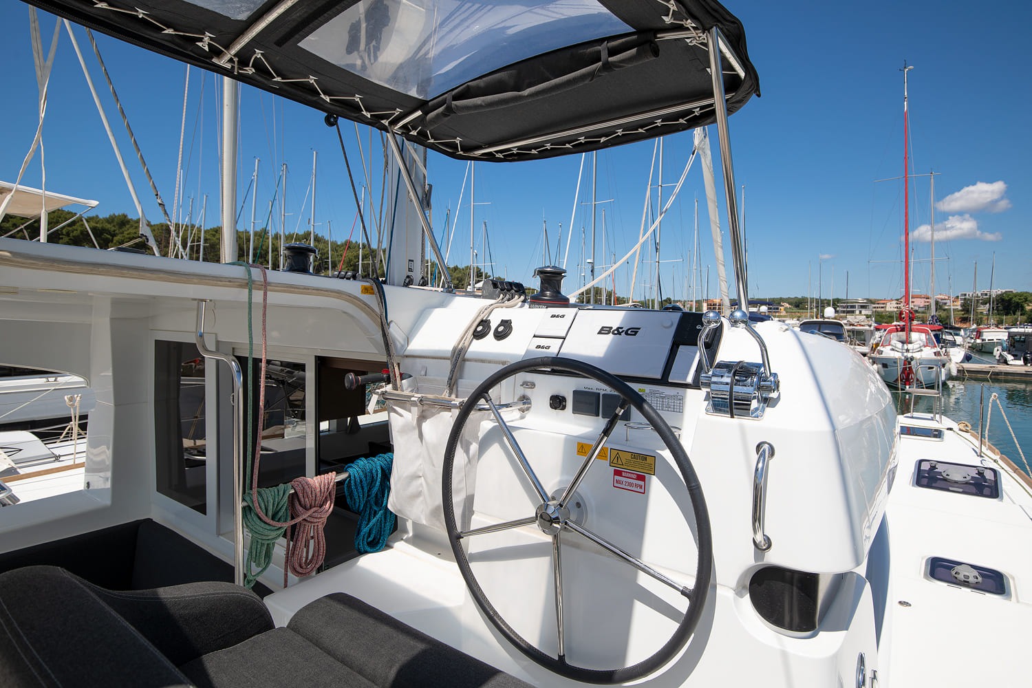 Yolo's raised starboard helm station with B&G instruments, winches and the wheel, with marina masts behind.