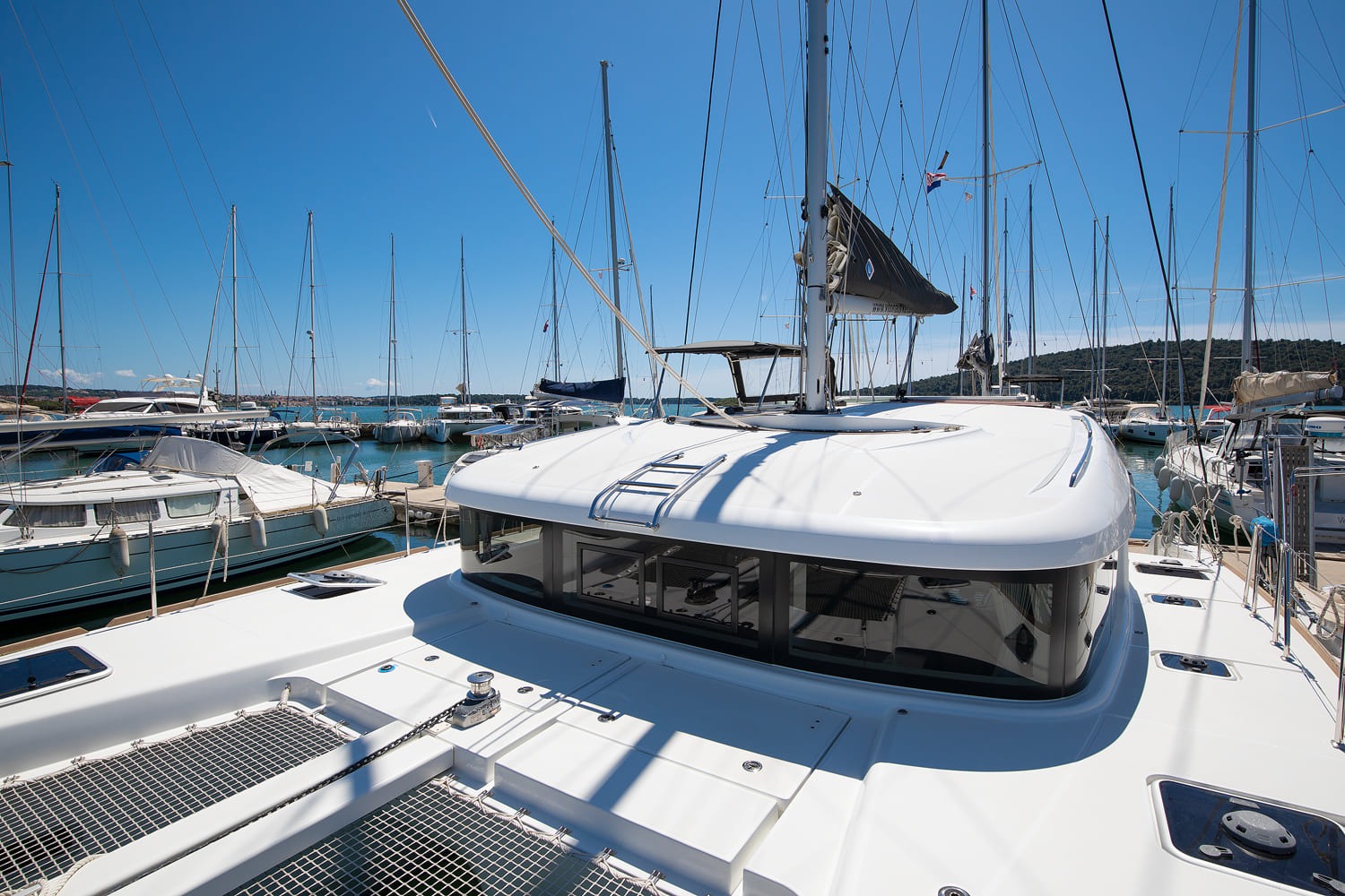 Foredeck and coachroof of the catamaran, with trampolines, anchor windlass and boats moored alongside in the marina.