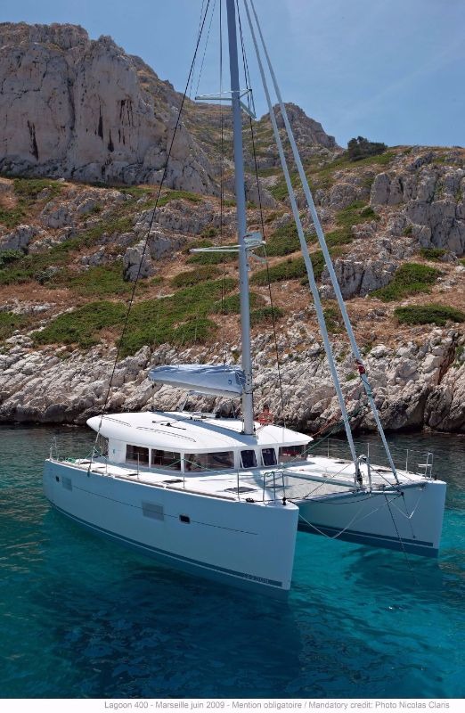 Lagoon 400 catamaran at anchor in clear turquoise water below a rocky cliff, seen from the bow with sails furled