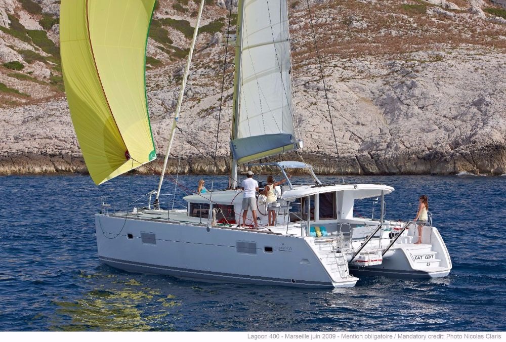 Crew on the deck of a Lagoon 400 catamaran sailing under a yellow gennaker along a rocky shore, with the helm station and bathing platform visible