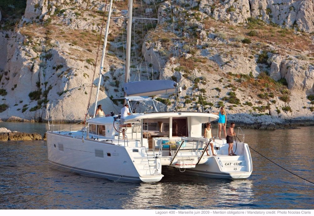 Lagoon 400 catamaran anchored below a rocky cliff, seen from astern with crew on both bathing platforms and the saloon doors open