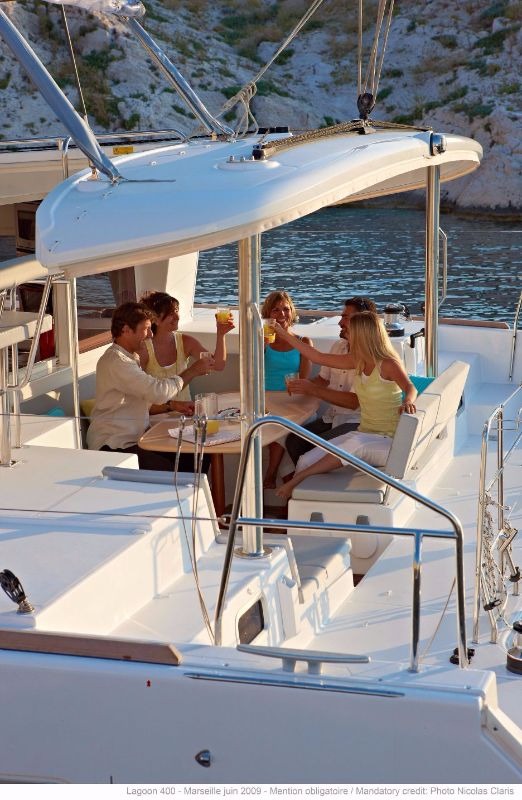 Six people seated around the table in the shaded cockpit of a Lagoon 400 catamaran, with the bathing platform and rocky shore behind