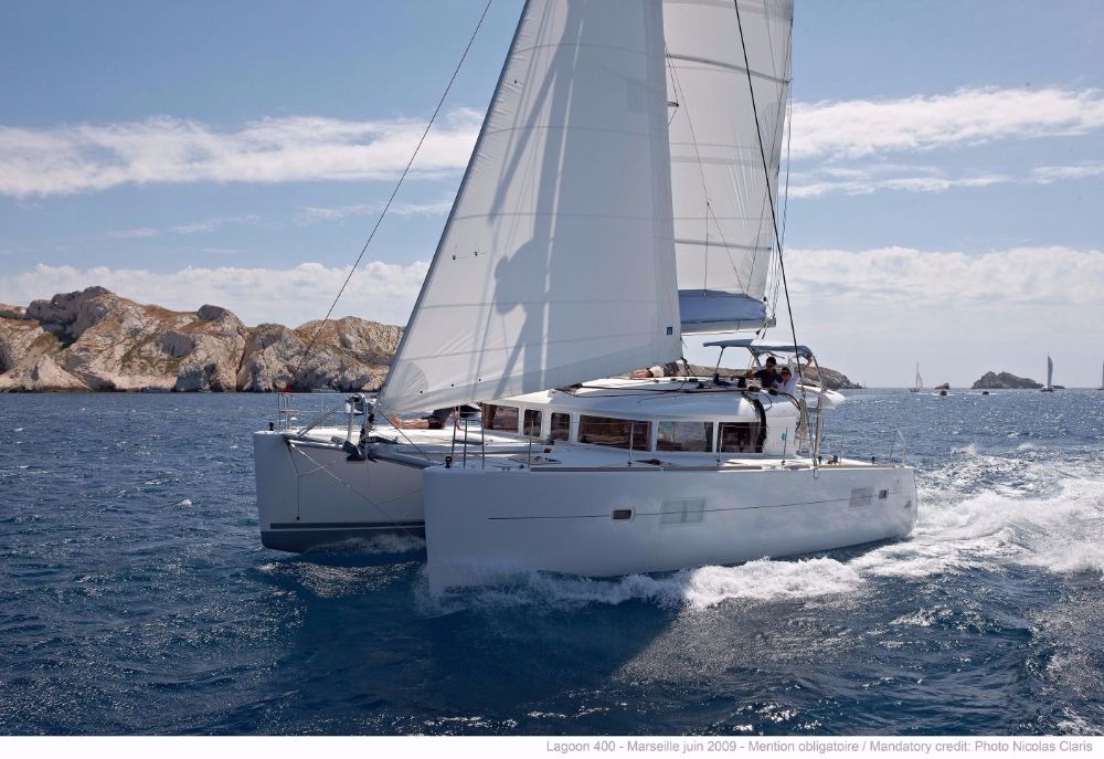 Lagoon 400 catamaran sailing upwind under full main and genoa, both bows throwing spray, with two people at the helm