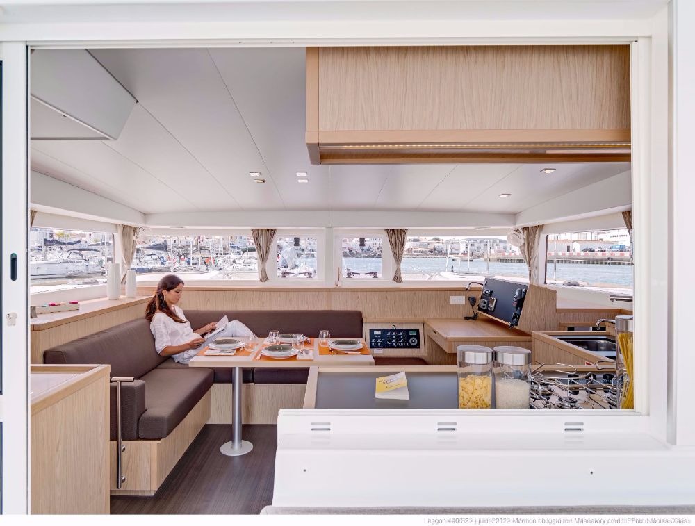 Saloon of a Lagoon 400 catamaran seen from the cockpit, with a woman reading at the dining table, the galley to the right and the navigation station beyond